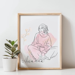 Puede incluir: Una impresión artística enmarcada con un dibujo lineal de una persona con un suéter rosa, sosteniendo una taza. La obra de arte incluye un lavado de acuarela y el nombre "Arianna" en cursiva. Una pequeña planta en maceta está a la izquierda del marco.