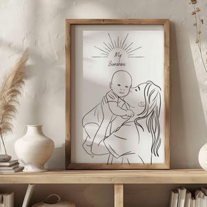 Puede incluir: Una impresión en blanco y negro enmarcada. La obra de arte representa a una madre sosteniendo a un bebé, con el texto "My Sunshine" arriba. El marco es de color marrón claro y la impresión se exhibe en una estantería de madera.