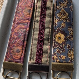 Puede incluir: Tres llaveros de tela para muñeca con herrajes metálicos. Los llaveros presentan diferentes diseños: un diseño floral naranja y morado, una tela verde con ribete burdeos y un estampado floral azul. Cada llavero tiene un anillo metálico.