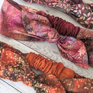 Könnte beinhalten: Drei Stoff-Stirnbänder mit einem Knotendesign. Die Stirnbänder sind aus einem weichen, dehnbaren Stoff mit einem Batikdruck in Braun-, Orange- und Rottönen.