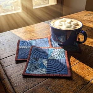 Könnte beinhalten: Zwei quadratische Stoffuntersetzer mit blauem Patchwork-Muster, kreisförmigen Steppnähten und einem dunkelroten Rand, platziert neben einer dunkelblauen Keramiktasse, die mit einem Kaffeegetränk und einer dicken Schicht weißem Milchschaum gefüllt ist.