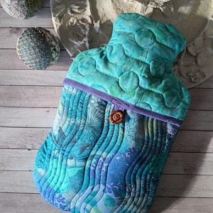 Könnte beinhalten: Eine gesteppte Wärmflaschenhülle in Türkis- und Blautönen mit floralem Muster und wellenförmigen Nähten. Sie hat eine lila Einfassung und einen Holzknopfverschluss. Auf einer Holzoberfläche.