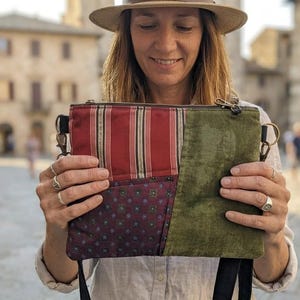Könnte beinhalten: Eine rechteckige Umhängetasche im Patchwork-Design mit einem rot-weiß gestreiften Bereich, einem dunkelburgunderfarbenen Feld mit kleinem geometrischen Muster und einem einfarbigen olivgrünen Abschnitt. Die Tasche hat einen Reißverschluss und Metallbeschläge.