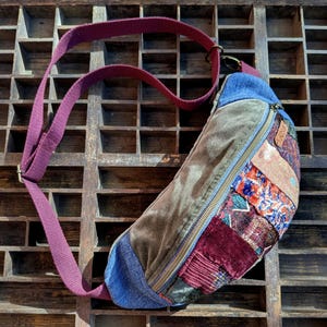 May include: A crossbody bag featuring a patchwork design with panels of olive green velvet, blue denim, and various patterned fabrics in shades of red, purple, and orange. The bag includes a long, adjustable maroon strap and a gold-toned metal zipper.
