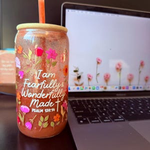 May include: A clear glass tumbler with a floral design and the text "I am Fearfully & Wonderfully Made" in white. The tumbler has a bamboo lid and an orange straw. The background includes a laptop with a floral wallpaper.