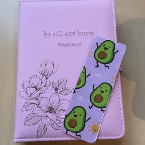 May include: A pink journal with the text "be still and know" and "PSALM 46:10" is next to a bookmark with avocado illustrations. The journal has a floral design. The bookmark features smiling avocados, clouds, and a sun.