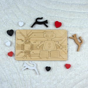 Pode incluir: Um quebra-cabeça de robô de madeira com detalhes de placa de circuito gravados e um design de robô sorridente. Formas de gato e coração de madeira em preto, branco e vermelho circundam o quebra-cabeça. Em uma superfície branca texturizada.