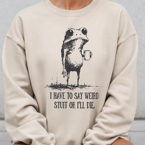 May include: Beige sweatshirt featuring a black and white illustration of a frog holding a mug. The text "I HAVE TO SAY WEIRD STUFF OR I'LL DIE" is printed below the frog. The sweatshirt has long sleeves and a crew neck.