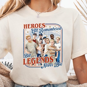 May include: Cream-colored t-shirt featuring a graphic of a group of boys with the text "HEROES Get Remembered But LEGENDS Never Die" in a blue and red design. The shirt is made of a soft material.