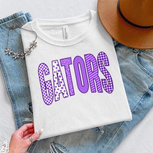 May include: A white t-shirt with the word "GATORS" in purple, featuring star and checkered patterns. The shirt is displayed on blue jeans, with a brown hat and a floral necklace.
