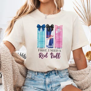 May include: Cream-colored t-shirt featuring a graphic of three Red Bull cans with bows, in blue, black, and pink. The text "First, I need a Red Bull" is printed below the cans in purple script. The shirt is worn by a person.