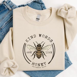 May include: A beige crewneck sweatshirt featuring a central graphic of a honeybee surrounded by a circular border. The text reads: KIND WORDS ARE LIKE HONEY PROVERBS 16:24. The sweatshirt is layered over a pair of blue denim jeans with visible seams.