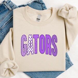May include: A beige crewneck sweatshirt featuring the word GATORS printed in large, purple-toned letters. Each letter displays a unique pattern, including checkerboard, stars, leopard print, grid, and diagonal stripes, all outlined in dark purple.