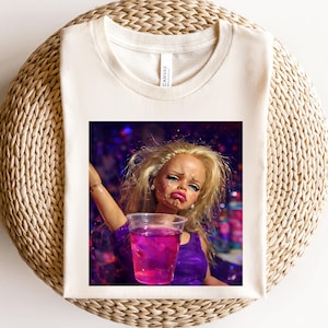 Könnte beinhalten: Cremefarbenes T-Shirt mit einer Grafik einer weinenden Puppe, die ein lila Getränk hält. Die Puppe hat blondes Haar, blauen Lidschatten und rosa Lippenstift. Das Bild hat einen Party-Hintergrund mit Konfetti.