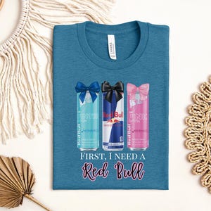May include: Teal t-shirt featuring three Red Bull cans with bows: blue, black, and pink. The text "FIRST, I NEED A Red Bull" is printed below the cans in a cursive font. The shirt has a crew neck.