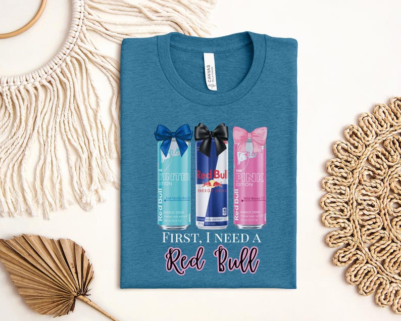 May include: Teal t-shirt featuring three Red Bull cans with bows: blue, black, and pink. The text "FIRST, I NEED A Red Bull" is printed below the cans in a cursive font. The shirt has a crew neck.