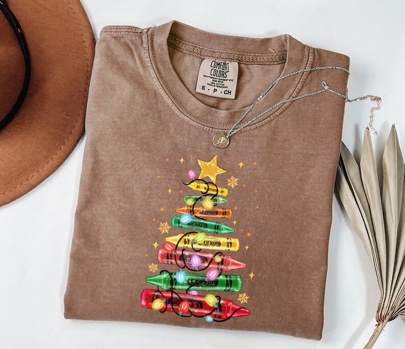 K&ouml;nnte beinhalten: Braunes Comfort Colors T-Shirt mit einem Weihnachtsbaum-Design aus Buntstiften. Der Baum besteht aus bunten Buntstiften, mit einem gelben Stern und Lichterketten. Eine silberne Halskette mit einem kleinen Anh&auml;nger liegt auf dem Shirt.