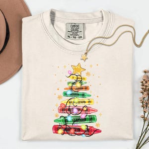 K&ouml;nnte beinhalten: Cremefarbenes Comfort Colors T-Shirt mit einem Weihnachtsbaum-Design aus Buntstiften. Der Baum ist mit Lichterketten und einem goldenen Stern geschm&uuml;ckt. Eine goldene Halskette mit Sternanh&auml;nger und ein brauner Hut sind ebenfalls im Bild.