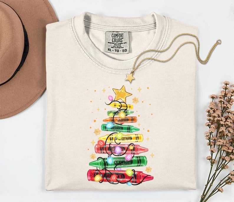 K&ouml;nnte beinhalten: Cremefarbenes Comfort Colors T-Shirt mit einem Weihnachtsbaum-Design aus Buntstiften. Der Baum ist mit Lichterketten und einem goldenen Stern geschm&uuml;ckt. Eine goldene Halskette mit Sternanh&auml;nger und ein brauner Hut sind ebenfalls im Bild.