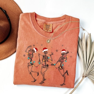 May include: A coral-colored Comfort Colors crew neck t-shirt featuring a graphic of three dancing skeletons wearing Santa hats and strung with Christmas lights. The shirt has a round neckline and short sleeves. A gold necklace is draped over the shirt.