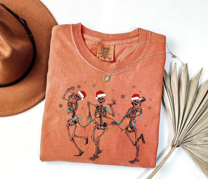 May include: A coral-colored Comfort Colors crew neck t-shirt featuring a graphic of three dancing skeletons wearing Santa hats and strung with Christmas lights. The shirt has a round neckline and short sleeves. A gold necklace is draped over the shirt.