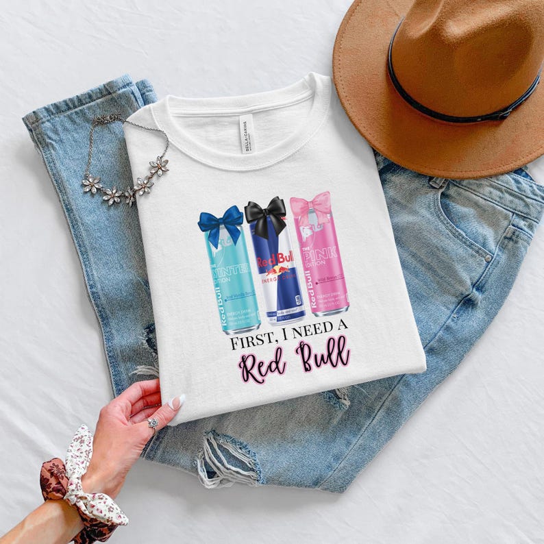 May include: White t-shirt featuring three Red Bull cans with bows, in blue, black, and pink. The text "First, I need a Red Bull" is printed below the cans. The shirt is laid on blue jeans with a brown hat.