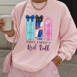 May include: Pink sweatshirt with a graphic of three Red Bull cans: Winter Edition, Energy Drink, and Pink Edition. The text "FIRST, I NEED A Red Bull" is printed below the cans. The sweatshirt is paired with pink striped shorts.