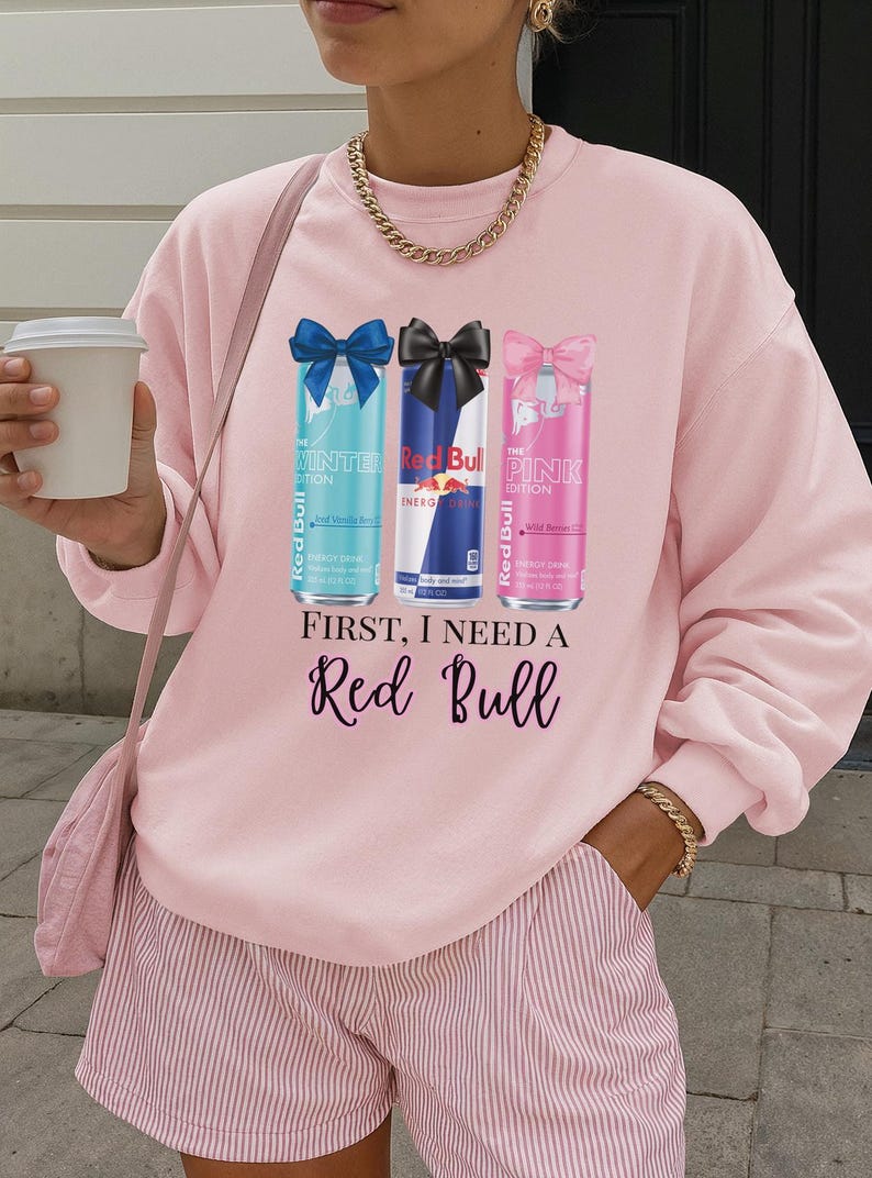 May include: Pink sweatshirt with a graphic of three Red Bull cans: Winter Edition, Energy Drink, and Pink Edition. The text "FIRST, I NEED A Red Bull" is printed below the cans. The sweatshirt is paired with pink striped shorts.