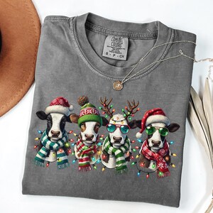 以下が含まれることがあります： クリスマス衣装を着た4頭の牛が描かれたグレーのTシャツ。各牛はサンタの帽子、トナカイの角、または冬の帽子をかぶり、スカーフとフェアリーライトを身に着けています。Tシャツはクルーネックで半袖です。