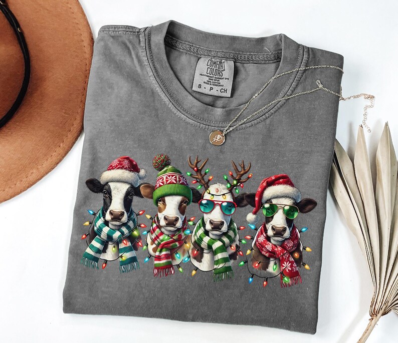 以下が含まれることがあります： クリスマス衣装を着た4頭の牛が描かれたグレーのTシャツ。各牛はサンタの帽子、トナカイの角、または冬の帽子をかぶり、スカーフとフェアリーライトを身に着けています。Tシャツはクルーネックで半袖です。