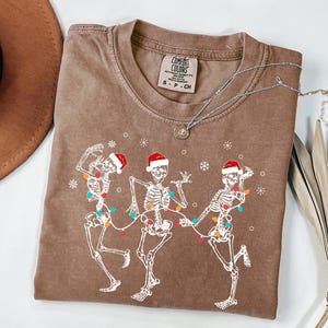 May include: Brown t-shirt featuring a graphic of three dancing skeletons wearing Santa hats and strung with Christmas lights. The shirt has a crew neck and a small tag at the top. A silver necklace with a pendant is draped across the shirt.
