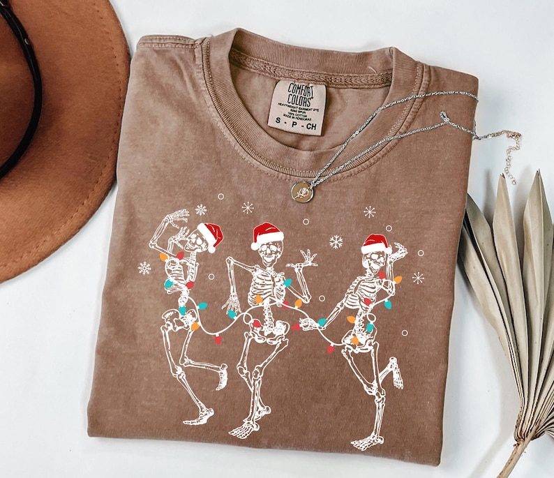 May include: Brown t-shirt featuring a graphic of three dancing skeletons wearing Santa hats and strung with Christmas lights. The shirt has a crew neck and a small tag at the top. A silver necklace with a pendant is draped across the shirt.