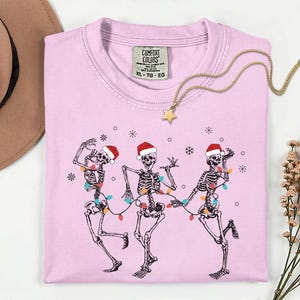 May include: A light pink t-shirt featuring a graphic of three dancing skeletons wearing Santa hats and strung with Christmas lights. A gold necklace with a star pendant rests on the shirt. The shirt has a Comfort Colors tag.