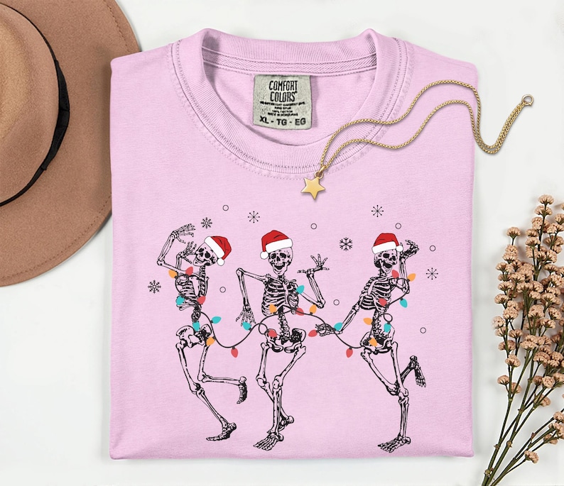 May include: A light pink t-shirt featuring a graphic of three dancing skeletons wearing Santa hats and strung with Christmas lights. A gold necklace with a star pendant rests on the shirt. The shirt has a Comfort Colors tag.