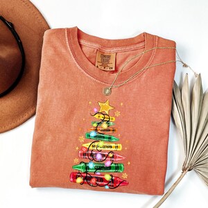 K&ouml;nnte beinhalten: Ein korallenfarbenes Comfort Colors Sweatshirt mit einem Weihnachtsbaum-Design aus bunten Buntstiften. Der Baum ist mit einem goldenen Stern und Lichterketten geschm&uuml;ckt. Eine goldene Halskette und ein brauner Hut sind ebenfalls im Bild.