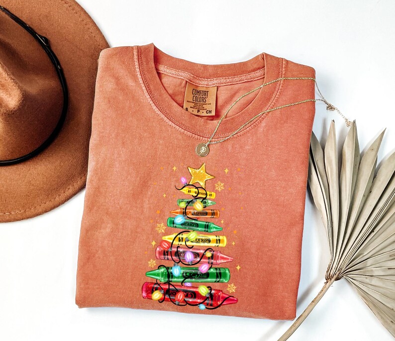K&ouml;nnte beinhalten: Ein korallenfarbenes Comfort Colors Sweatshirt mit einem Weihnachtsbaum-Design aus bunten Buntstiften. Der Baum ist mit einem goldenen Stern und Lichterketten geschm&uuml;ckt. Eine goldene Halskette und ein brauner Hut sind ebenfalls im Bild.