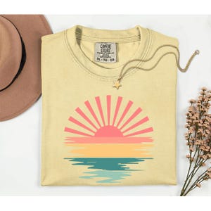 May include: A light yellow Comfort Colors t-shirt with a sunset graphic in pink, orange, yellow, and teal. The shirt has a crew neck and short sleeves. A gold necklace with a star pendant and a brown hat are next to the shirt.