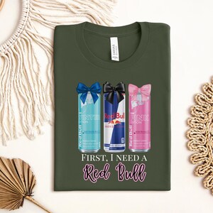 May include: An olive green t-shirt featuring three Red Bull cans with bows. The cans are the Winter, Original, and Pink editions. The text "FIRST, I NEED A Red Bull" is below the cans. The shirt has a crew neck.