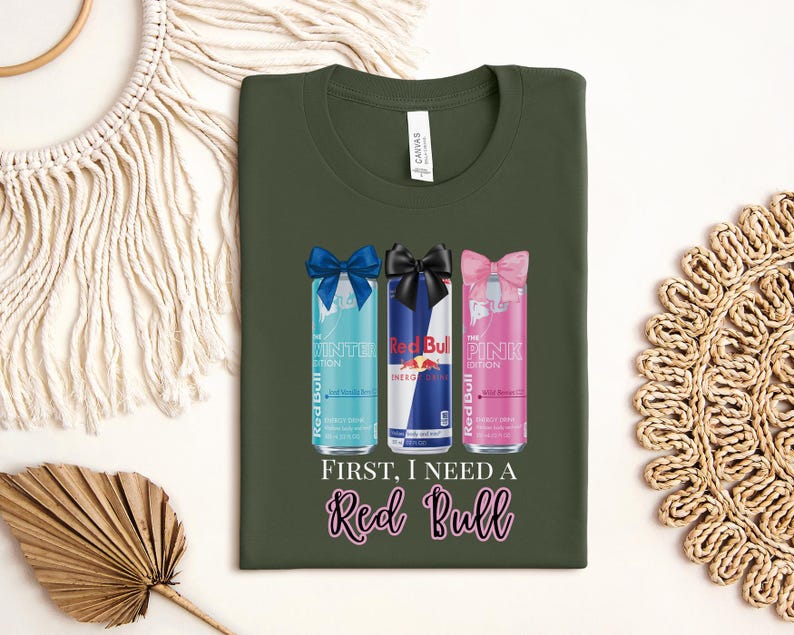 May include: An olive green t-shirt featuring three Red Bull cans with bows. The cans are the Winter, Original, and Pink editions. The text "FIRST, I NEED A Red Bull" is below the cans. The shirt has a crew neck.