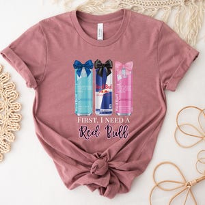May include: Mauve-colored t-shirt featuring a graphic of three Red Bull cans with bows. The cans are blue, red, and pink. Text on the shirt reads "First, I need a Red Bull" in a cursive font. The shirt is knotted at the bottom.