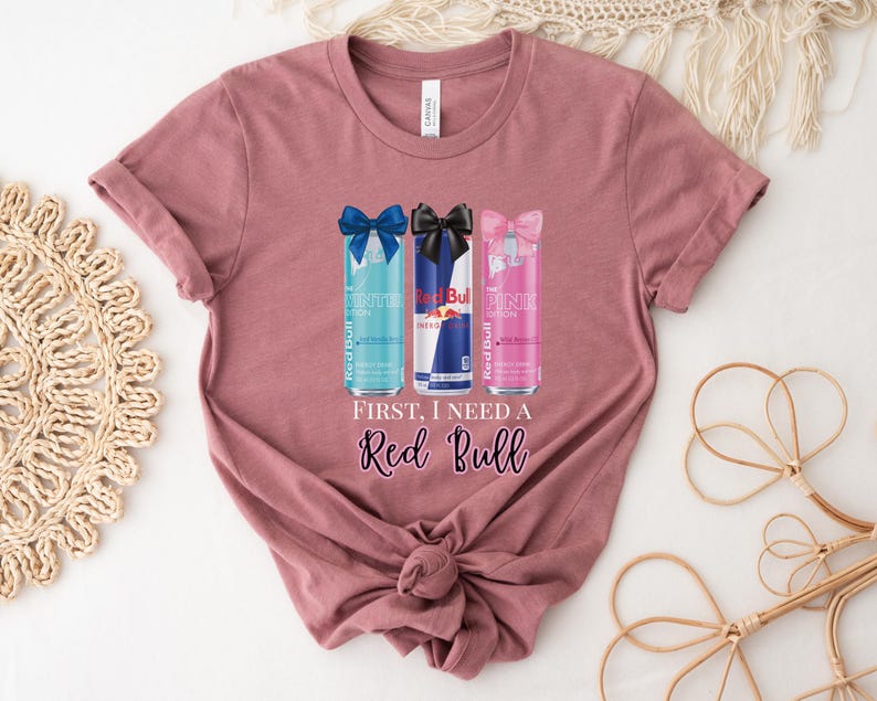 May include: Mauve-colored t-shirt featuring a graphic of three Red Bull cans with bows. The cans are blue, red, and pink. Text on the shirt reads "First, I need a Red Bull" in a cursive font. The shirt is knotted at the bottom.