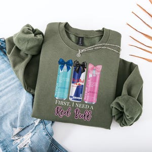 May include: An olive green sweatshirt featuring three Red Bull can designs with bows, and the text "First, I need a Red Bull" in pink script. A silver necklace and a pair of blue jeans are also in the image.