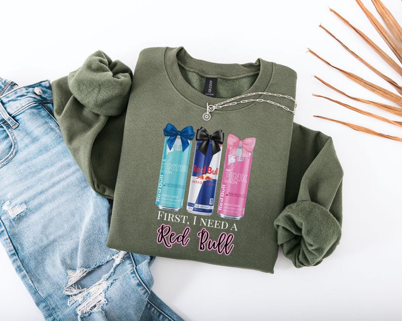May include: An olive green sweatshirt featuring three Red Bull can designs with bows, and the text "First, I need a Red Bull" in pink script. A silver necklace and a pair of blue jeans are also in the image.