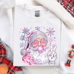 Könnte beinhalten: Weißes Sweatshirt mit Rundhalsausschnitt und Santa Claus-Grafik. Santa trägt eine Sonnenbrille, einen Discokugelhut und bläst eine Kaugummiblase. Das Design umfasst rosa und lila Schneeflocken und ein Friedenszeichen.