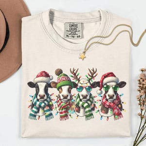 以下が含まれることがあります： クリーム色のComfort Colors XL-TG-EG Tシャツ。お祝いの牛のデザインが特徴です。サンタ帽、トナカイの角、サングラスを身に着けた4頭の牛が、カラフルな電飾とスカーフで飾られています。金の星のネックレスがシャツの上にあります。