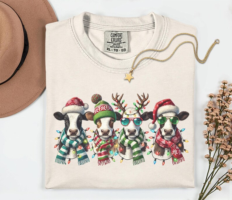 以下が含まれることがあります： クリーム色のComfort Colors XL-TG-EG Tシャツ。お祝いの牛のデザインが特徴です。サンタ帽、トナカイの角、サングラスを身に着けた4頭の牛が、カラフルな電飾とスカーフで飾られています。金の星のネックレスがシャツの上にあります。