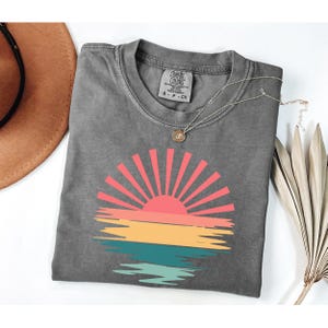 May include: A gray Comfort Colors t-shirt featuring a colorful sunset design. The sunset graphic includes a red and orange sun with yellow and teal water. A silver necklace with a small pendant is on the shirt.