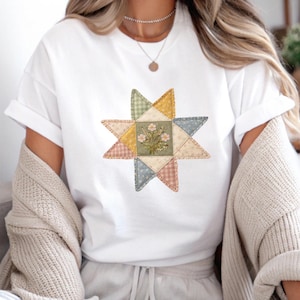 Puede incluir: Una camiseta blanca de manga corta con un gráfico central de una estrella de múltiples puntas compuesta por varios parches de tela estampada en tonos apagados de amarillo, verde, azul y rosa, con un pequeño ramo de flores bordado en el centro.