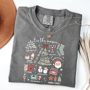 Könnte beinhalten: Graues Comfort Colors T-Shirt mit einem festlichen Weihnachtsgrafik. Das Design umfasst einen Weihnachtsbaum, den Weihnachtsmann, Zuckerstangen und den Satz "tis the season". Eine goldene Halskette und ein brauner Hut sind ebenfalls sichtbar.