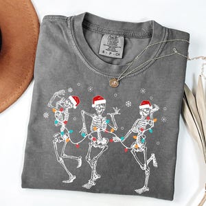 May include: Gray t-shirt featuring a graphic of three dancing skeletons wearing Santa hats and strung with Christmas lights. The skeletons are white, and the lights are colorful. The shirt has a crew neck and short sleeves.